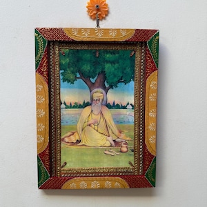 Puede incluir: Una obra de arte religiosa enmarcada representa una figura con túnicas amarillas sentada bajo un árbol. La obra de arte tiene un marco decorativo con detalles en rojo, verde y dorado. La imagen está colgada en una pared blanca con un gancho en forma de flor naranja.