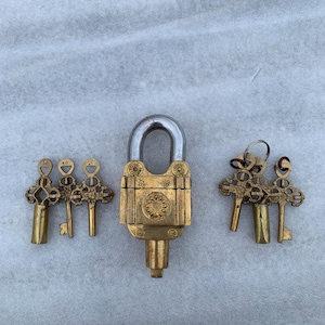 May include: A brass padlock with intricate details and five matching keys. The padlock has a silver-colored shackle and a decorative design on the front. The keys are ornate and have a similar design to the padlock.
