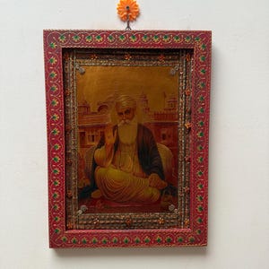May include: A framed religious artwork featuring a seated figure with a long white beard and a raised hand. The artwork has a gold background and is set within a red frame with decorative detailing. An orange flower-shaped hook is above the frame.