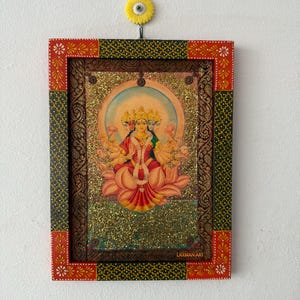 May include: A framed artwork depicting a multi-armed deity seated on a lotus flower, with a golden halo. The frame is red, green, and gold, with floral and geometric patterns. The artwork is labelled "LAXMAN ART".