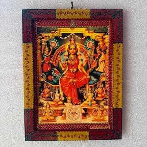 Puede incluir: Una impresión religiosa enmarcada que presenta una deidad central con vestimenta roja, rodeada de otras figuras. La obra de arte está dentro de un marco decorativo con detalles en rojo, amarillo y dorado. La impresión incluye el texto "LAXMAN ART".