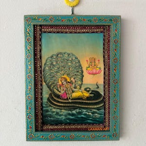 Könnte beinhalten: Ein gerahmtes religiöses Kunstwerk mit einer Gottheit, die auf einer Schlange liegt, mit einem türkis-goldenen Rahmen. Das Kunstwerk zeigt eine Figur in gelben und rosa Gewändern, umgeben von Wasser und einer großen Schlange. Der Rahmen hat Blumen- und Juwelenakzente.