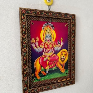May include: A framed artwork depicting a deity riding a lion, set against a vibrant red and purple background. The deity is adorned in red and gold, with multiple arms holding various objects. The frame is brown with floral and geometric patterns.