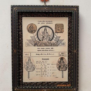 May include: Framed vintage print with religious figures and text in a decorative black frame. The print features illustrations of deities and script. An orange flower-shaped hook is above the frame.