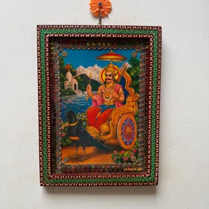 Op de afbeelding: Een religieuze prent in een lijst met een afbeelding van een godheid in een strijdwagen, met een zwarte kraai op de voorgrond. De lijst is versierd met groene en rode accenten. De prent hangt aan een witte muur met een oranje bloemvormige haak.