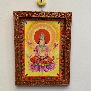Può includere: Una stampa religiosa incorniciata raffigurante una divinità seduta con più braccia, ornata di gioielli e una corona, circondata da un'esplosione solare. L'opera d'arte è racchiusa in una cornice marrone decorativa con accenti floreali, appesa a una parete bianca.