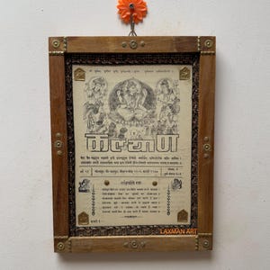 May include: A framed print with a religious theme. The wooden frame has decorative brass accents. The print features illustrations and text in a foreign language. An orange flower-shaped hook is above the frame.