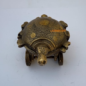 May include: A small, ornate brass turtle figurine with wheels. The turtle's shell is decorated with intricate patterns and gold accents. The turtle is on a white background. "LAXMAN ART" is printed on the turtle's shell.