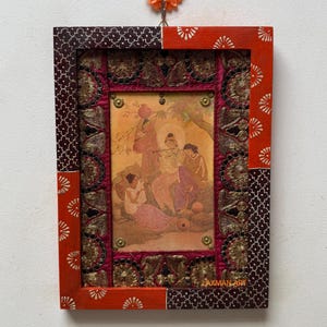 May include: A framed artwork with a traditional Indian painting. The frame is a patchwork of red, patterned brown, and gold fabric. The artwork depicts figures in a landscape. The text "LAXMAN-ART" is visible on the bottom right corner.