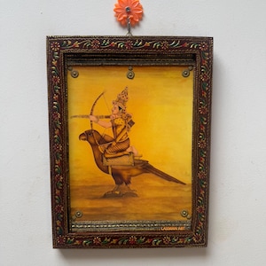 Könnte beinhalten: Ein verzierter, gerahmter Gemälde zeigt eine Figur mit Pfeil und Bogen auf einem großen Vogel vor gelbem Hintergrund. Der dunkelbraune Rahmen hat florale und goldene Akzente. Der Text "LAXMAN ART" ist sichtbar.