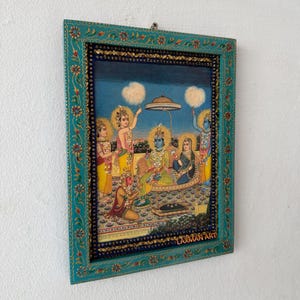 Pode incluir: Uma obra de arte emoldurada retratando uma cena religiosa com figuras em roupas coloridas. A moldura turquesa tem padrões florais e geométricos. A obra apresenta uma figura central com pele azul, cercada por outras figuras e elementos decorativos. O texto "LAXMAN ART" é visível.