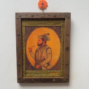 May include: A framed portrait of a man in traditional attire, set against an orange background. The wooden frame is adorned with decorative metal accents and a gold border. The artwork is hanging on a white wall.
