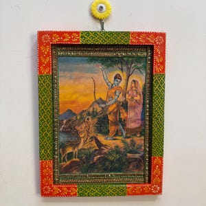 Op de afbeelding: Een religieus schilderij in een lijst met figuren in traditionele kledij met pijl en boog, tegen een landschap. De lijst is rood en groen met bloemmotieven en wordt opgehangen met een gele bloemvormige haak.
