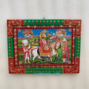 May include: A framed painting depicting a religious figure riding a white horse. The painting is surrounded by a green and red frame with gold accents. The text "LAXMAN - ART" is visible at the bottom of the frame.