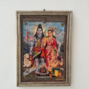 May include: A framed religious print featuring Hindu deities. The central figures are a male deity with blue skin, holding a trident, and a female deity in a red sari. A small elephant-headed deity kneels nearby. The frame is a light brown color.