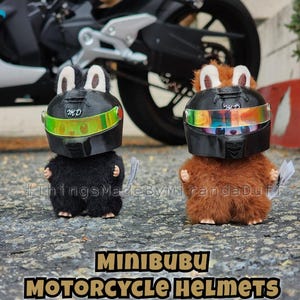 Könnte beinhalten: Zwei Plüschtierfiguren mit Miniatur-Motorradhelmen. Eine Figur ist schwarz, die andere braun. Die Helme sind schwarz mit schillernden Visieren. Der Text "MINIBUBU MOTORCYCLE HELMETS" ist darunter abgebildet.