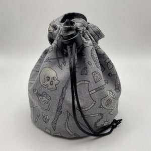 May include: Gray fabric dice bag with a drawstring closure. The bag is decorated with a repeating pattern of black and white illustrations of dice, skulls, swords, and other fantasy-themed imagery.