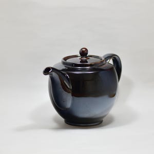 May include: A dark brown, glossy ceramic teapot with a rounded body, a curved spout, a matching lid with a small knob handle, and a side handle. The surface features a smooth, reflective finish with subtle variations in the deep brown glaze.