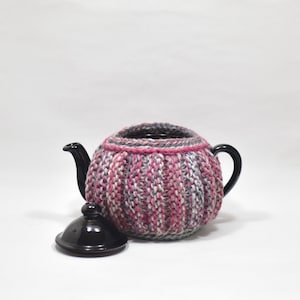 May include: A black ceramic teapot covered in a thick, ribbed, hand-knitted cozy featuring a variegated yarn pattern in shades of pink, grey, and white. The teapot has a matching black ceramic lid placed separately in front of the main body.