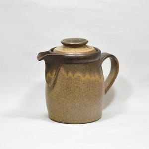 May include: A ceramic teapot with a speckled tan and brown glaze. The body features a rounded shape with a curved handle and a distinct spout. A matching lid with a central knob sits on top. The glaze transitions from a lighter tan base to a darker brown drip pattern near the rim and spout, creating a textured appearance.