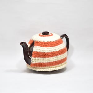 May include: A dark brown ceramic teapot covered by a crocheted tea cozy featuring horizontal stripes in cream and burnt orange. The cozy fits snugly around the body of the teapot, leaving the dark brown spout and handle exposed for functional use.