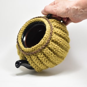 May include: A green knitted teapot cosy with a brown trim. The cosy is covering a black teapot.