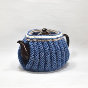 May include: A dark brown ceramic teapot encased in a thick, ribbed, blue knitted cozy. The cozy features a decorative band around the top rim with horizontal stripes in white, light gray, and dark blue, leaving the spout and handle exposed.