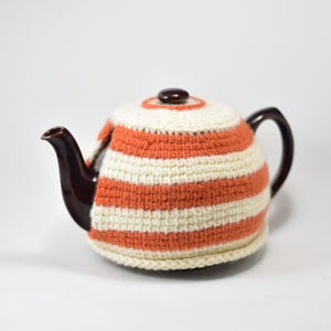 May include: A white and orange striped crocheted teapot cozy. The cozy is made of yarn and has a white crocheted border.