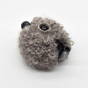 May include: A black ceramic teapot encased in a thick, textured, grey wool cozy. The cozy features a dense, curly pile and covers the body of the teapot, leaving the black spout and handle visible, with a circular opening at the top for the lid.