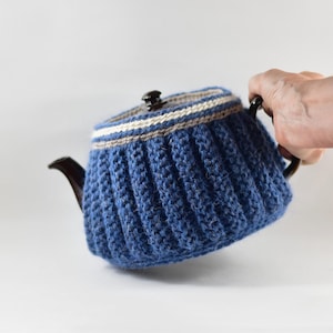 May include: A blue knitted tea cozy with a ribbed design, covering a dark brown teapot. The cozy has a striped band of beige and white near the top. The teapot has a dark brown handle, spout, and lid.