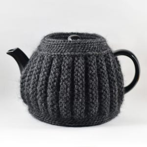 May include: A dark grey teapot with a knitted cosy. The cosy has a ribbed design and covers the body of the teapot. The teapot has a black handle, spout, and lid.