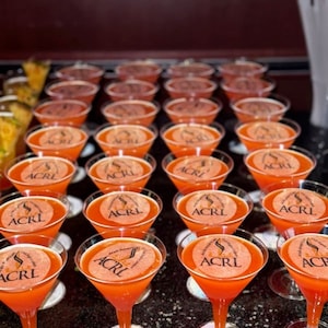 Puede incluir: Una fila de copas de martini de vidrio transparente llenas de un l&iacute;quido naranja brillante. Cada copa tiene una pegatina redonda con el texto "ACRL" en negro.