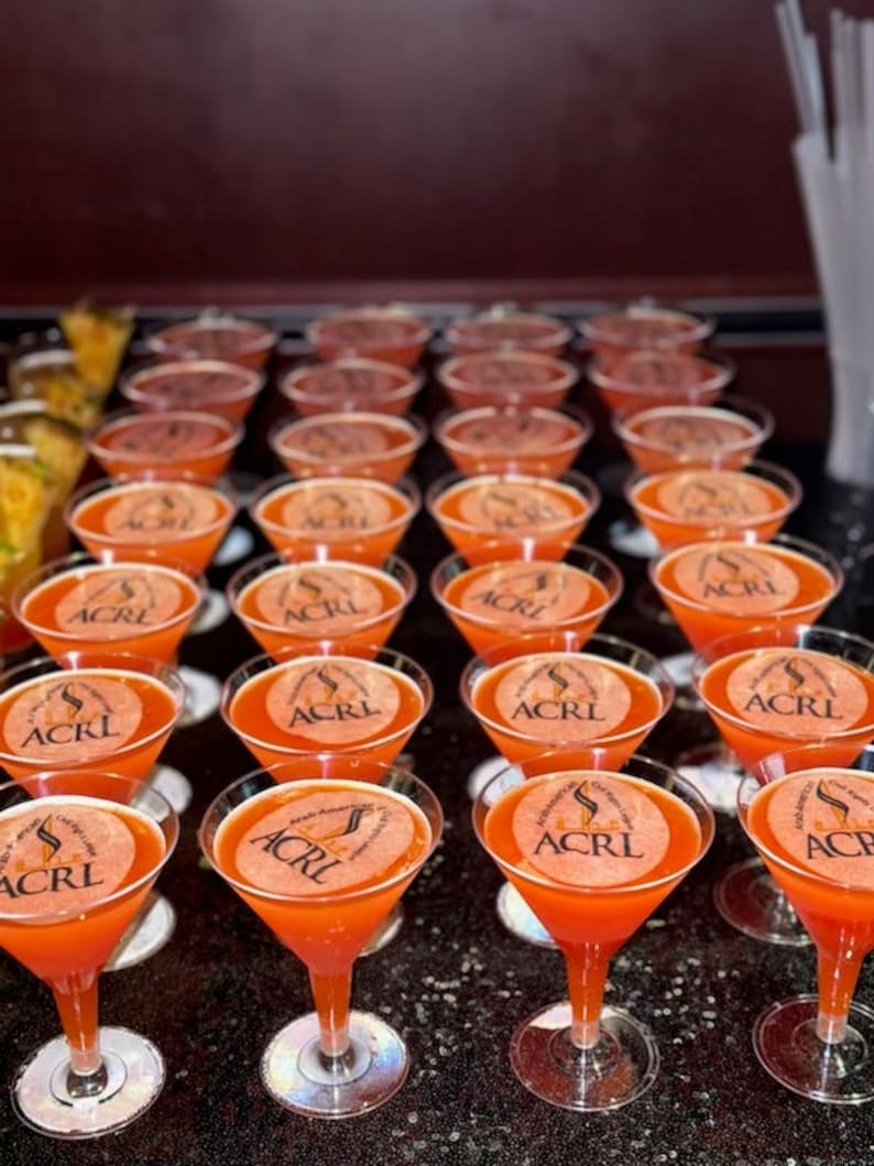 Puede incluir: Una fila de copas de martini de vidrio transparente llenas de un l&iacute;quido naranja brillante. Cada copa tiene una pegatina redonda con el texto "ACRL" en negro.