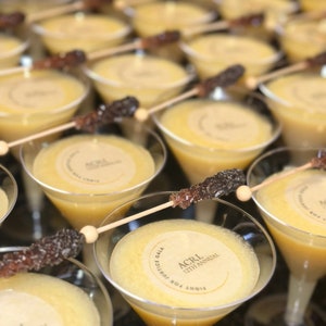Puede incluir: Primer plano de una mesa con m&uacute;ltiples copas de martini llenas de un l&iacute;quido amarillo. Cada copa tiene una pegatina redonda de papel con el texto "Fight For Justice Gala, ACRL 12th Annual".