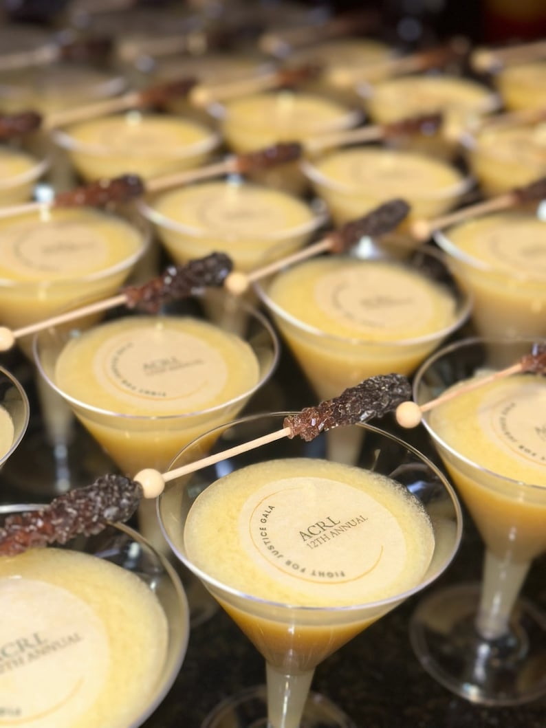 Puede incluir: Primer plano de una mesa con m&uacute;ltiples copas de martini llenas de un l&iacute;quido amarillo. Cada copa tiene una pegatina redonda de papel con el texto "Fight For Justice Gala, ACRL 12th Annual".