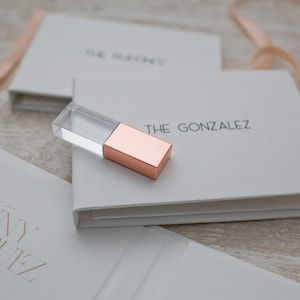 May include: Two white boxes with the names "The Gonzalez" and "Jenny Rodriguez" printed on them. A clear glass and rose gold USB drive is on top of the box with "The Gonzalez" printed on it.
