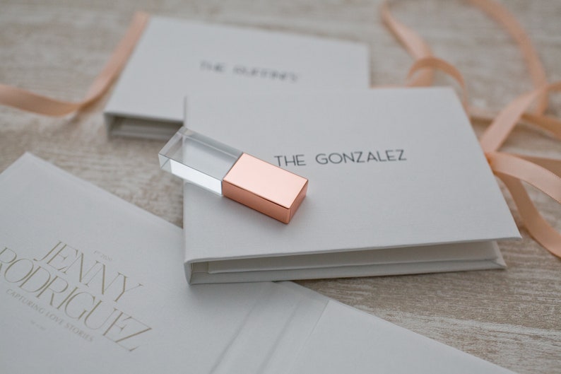 May include: Two white boxes with the names "The Gonzalez" and "Jenny Rodriguez" printed on them. A clear glass and rose gold USB drive is on top of the box with "The Gonzalez" printed on it.