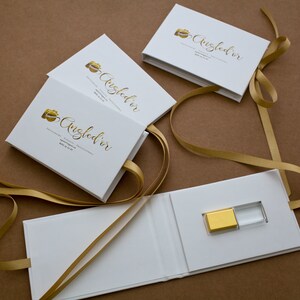 K&ouml;nnte beinhalten: Wei&szlig;e Geschenkboxen mit goldener Schleife und einem goldenen und durchsichtigen USB-Stick. Die Boxen tragen den Text "Angled'or" in goldenen Buchstaben und die Worte "Fotografie, Videografie, Professionalit&auml;t" in kleineren schwarzen Buchstaben.