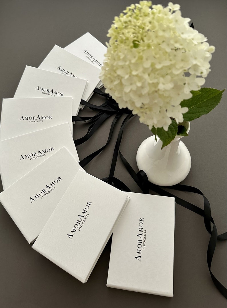K&ouml;nnte beinhalten: Ein Stapel wei&szlig;er Boxen mit schwarzem Text, der auf jeder Box "AMOR AMOR FOTOGRAFIA" lautet. Die Boxen sind mit schwarzem Band zusammengebunden und eine wei&szlig;e Blume steht in einer wei&szlig;en Vase auf einer grauen Oberfl&auml;che.