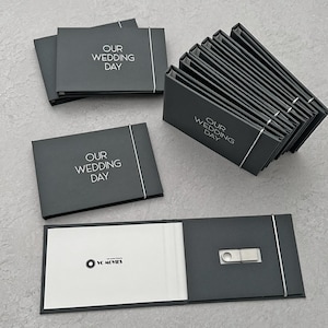 Puede incluir: Una pila de cajas de cartulina negra con una cinta blanca y el texto "Our Wedding Day" impreso en la parte delantera. Las cajas están dispuestas en forma de pirámide. La caja superior está abierta y revela una unidad USB plateada.