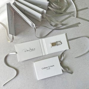Op de afbeelding: Witte rechthoekige kartonnen mappen met zilveren linten. Een geopende map toont een zilveren USB-stick en de tekst Robin Ganter Photography. Een gesloten map bevat de gedrukte tekst Colleen & Mark 9.5.26 in zwarte inkt.