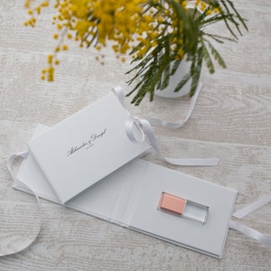 May include: White gift box with a white ribbon and the text "Alexandra & Daniel" printed on the front. The box is open to reveal a small, pink and clear USB drive.