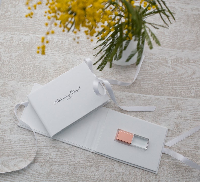 May include: White gift box with a white ribbon and the text "Alexandra & Daniel" printed on the front. The box is open to reveal a small, pink and clear USB drive.