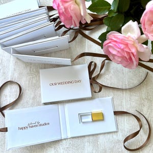 K&ouml;nnte beinhalten: Eine wei&szlig;e Box mit einem goldenen USB-Stick im Inneren. Die Box ist ge&ouml;ffnet und mit einem braunen Band umwickelt. Auf der Vorderseite der Box steht "OUR WEDDING DAY" und innen "filmed by happy haven studios".