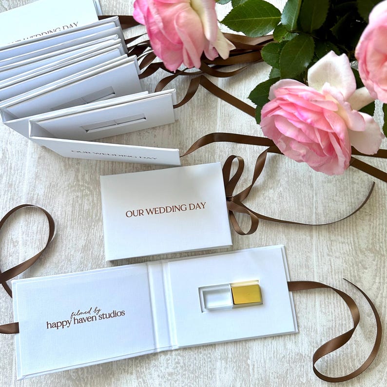 K&ouml;nnte beinhalten: Eine wei&szlig;e Box mit einem goldenen USB-Stick im Inneren. Die Box ist ge&ouml;ffnet und mit einem braunen Band umwickelt. Auf der Vorderseite der Box steht "OUR WEDDING DAY" und innen "filmed by happy haven studios".
