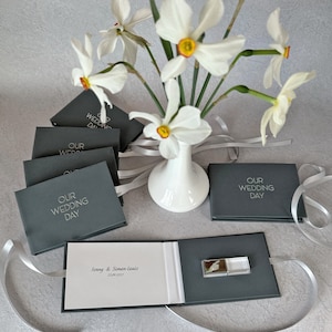 May include: A gray wedding favor box with a white ribbon and the text "Our Wedding Day" on the front. The box is open to reveal a small white book with the text "Jenny & Simon-Louis 23.29.2023" on the inside. A white USB drive is visible in the box.