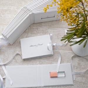 May include: White photo album with a white ribbon and a USB drive inside. The album has the text "Alexandra & Daniel" on the cover.