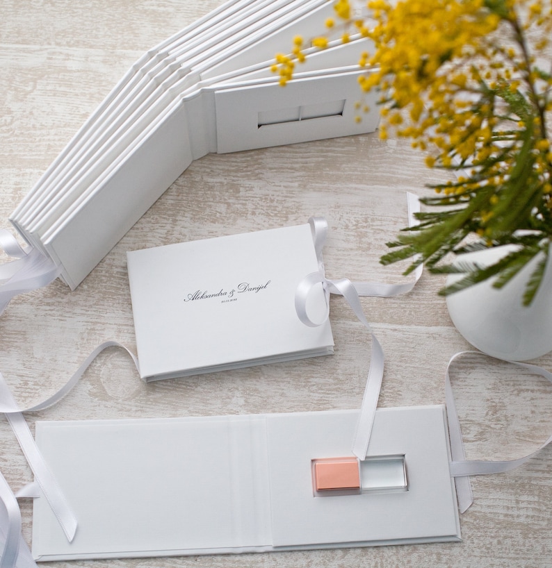 May include: White photo album with a white ribbon and a USB drive inside. The album has the text "Alexandra & Daniel" on the cover.