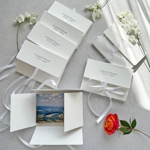 May include: White cardstock photo holders with the text "A Quick Glimpse XOXO, Tabitha" printed in black. Each holder is tied with a white ribbon. One holder is open, revealing a landscape photo. A red and orange rose and white flowers are also present.