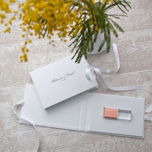 May include: White gift box with a white ribbon and a small pink and clear USB drive inside. The box has the text "Alexandra & Daniel" printed on the top.